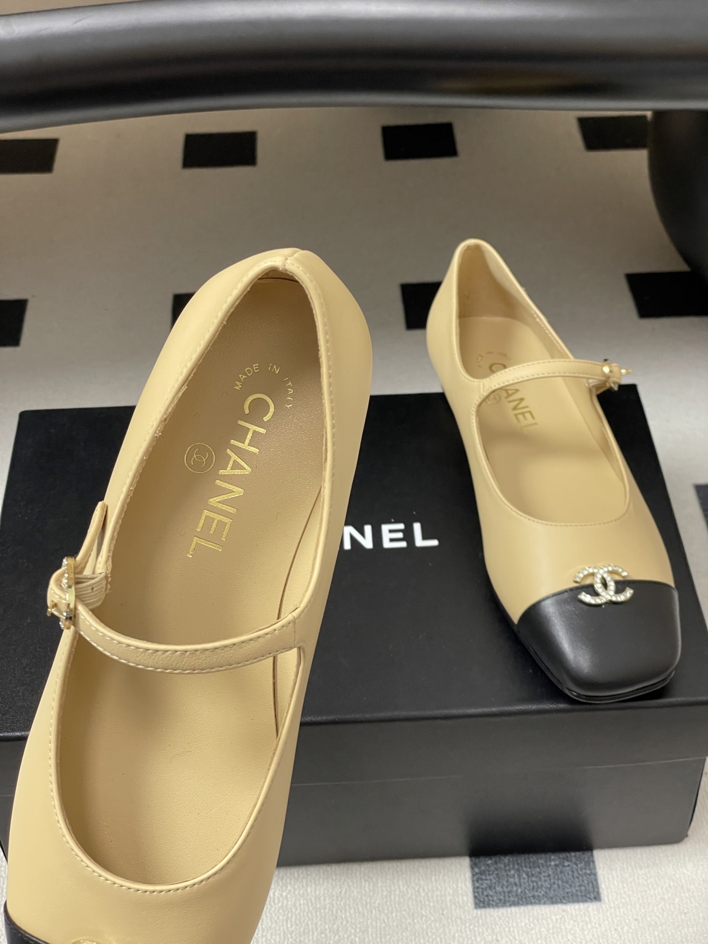 Pair of beige and black Chanel Mary Jane flats with crystal CC logos and rhinestone buckles on a black table - Hình 7