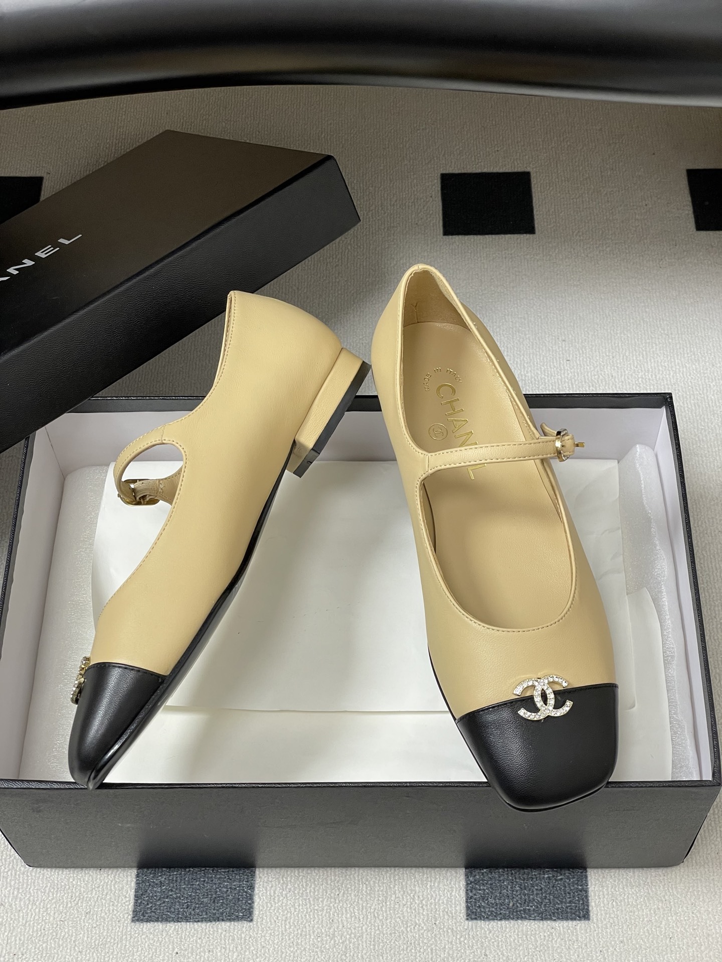 Pair of beige and black Chanel Mary Jane flats with crystal CC logos and rhinestone buckles on a black table - Hình 6