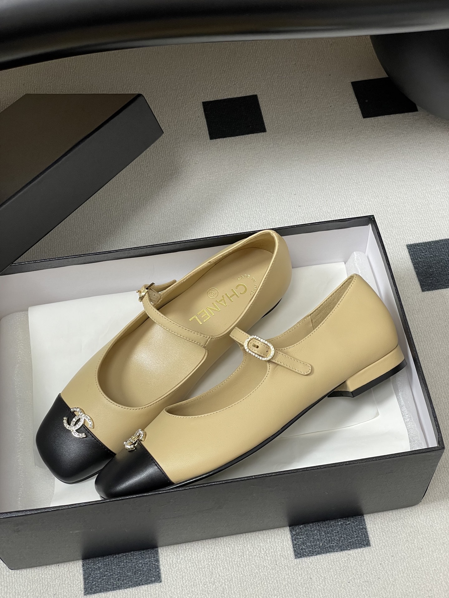 Pair of beige and black Chanel Mary Jane flats with crystal CC logos and rhinestone buckles on a black table - Hình 4
