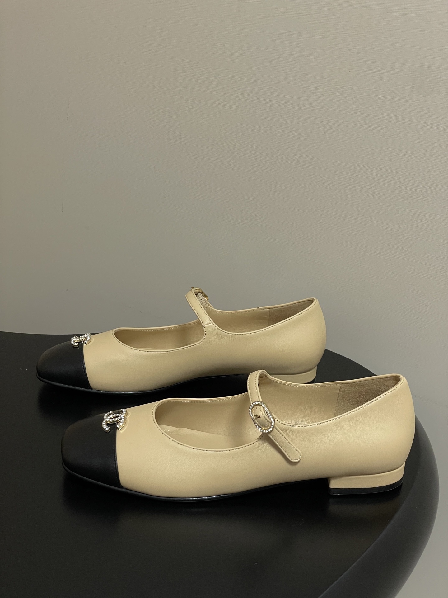 Pair of beige and black Chanel Mary Jane flats with crystal CC logos and rhinestone buckles on a black table - Hình 3