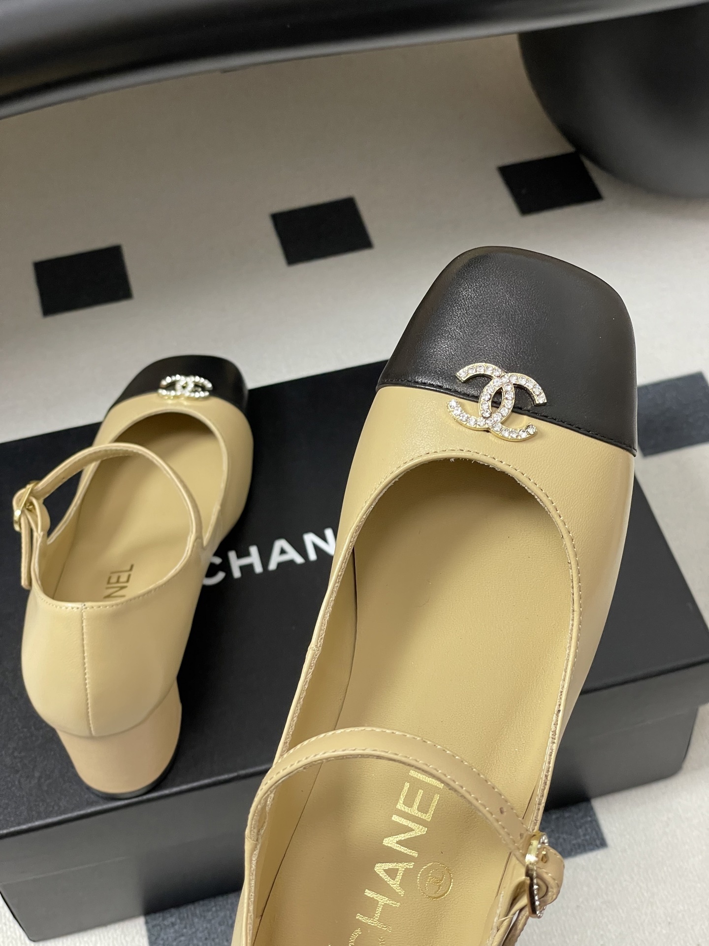 A pair of beige and black two-tone Mary Jane block heel shoes with crystal CC logos on a black table. - Hình 9