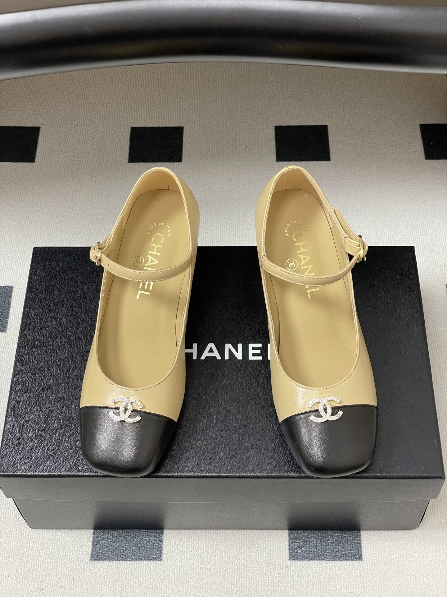 A pair of beige and black two-tone Mary Jane block heel shoes with crystal CC logos on a black table. - Hình 5