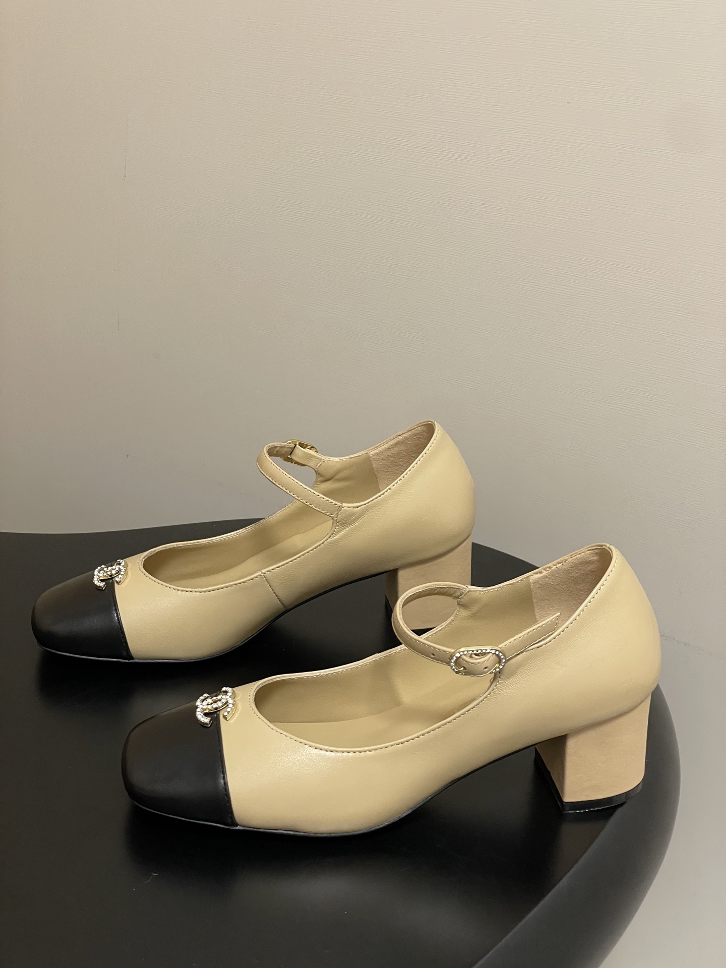 A pair of beige and black two-tone Mary Jane block heel shoes with crystal CC logos on a black table. - Hình 3