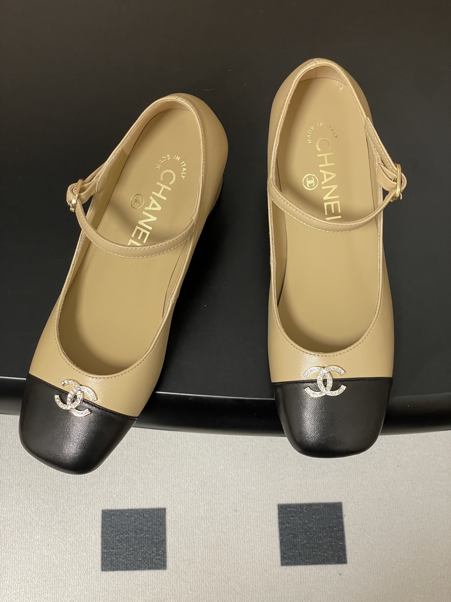 A pair of beige and black two-tone Mary Jane block heel shoes with crystal CC logos on a black table. - Hình 2