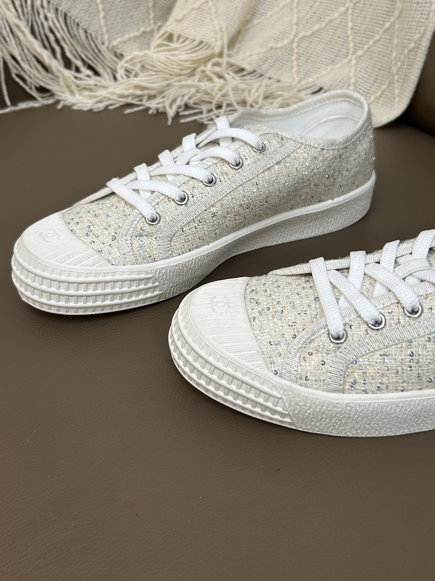 A pair of Chanel beige tweed low-top sneakers with sparkle details and white laces on a brown leather surface. - Hình 9