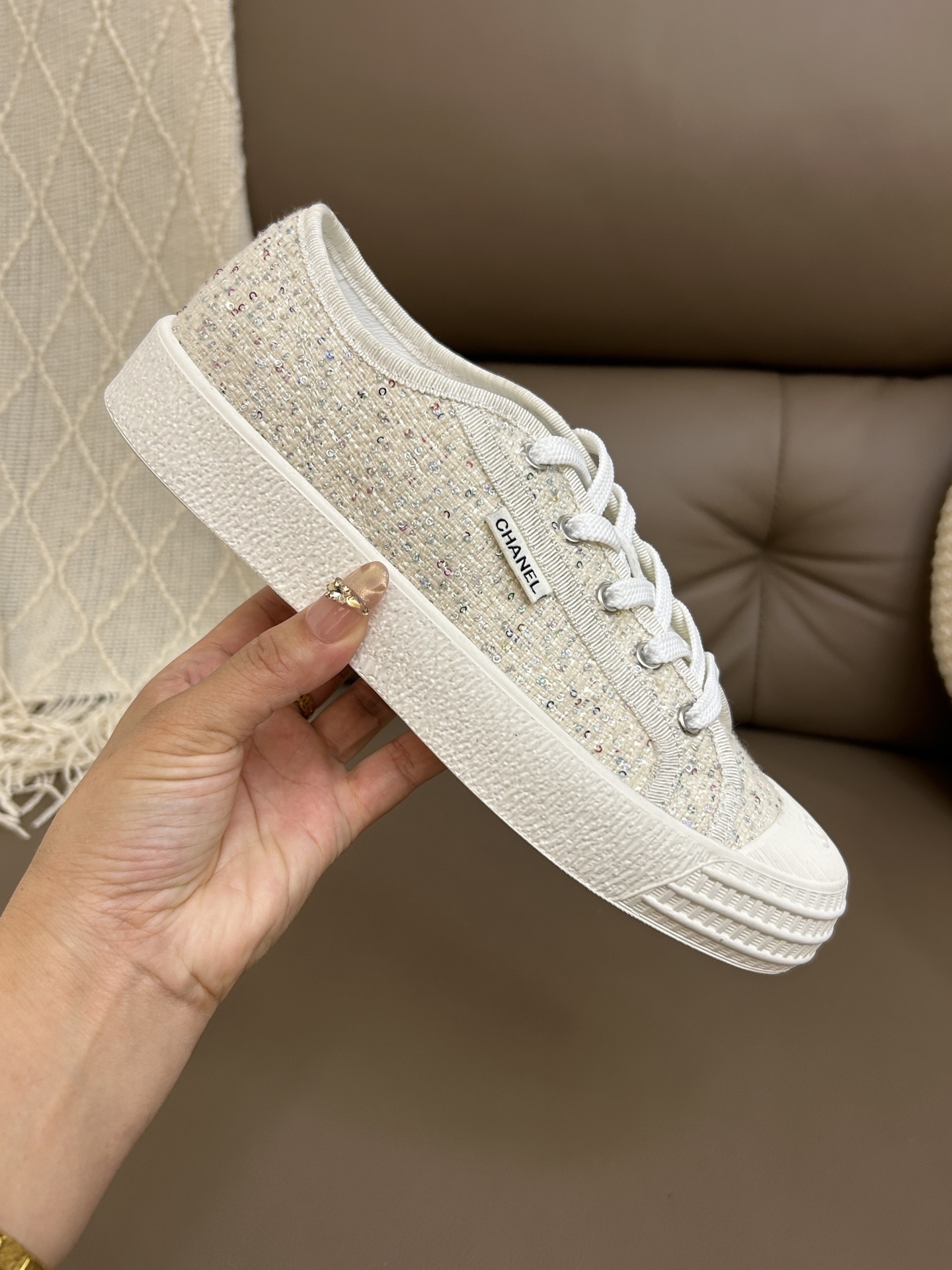 A pair of Chanel beige tweed low-top sneakers with sparkle details and white laces on a brown leather surface. - Hình 8