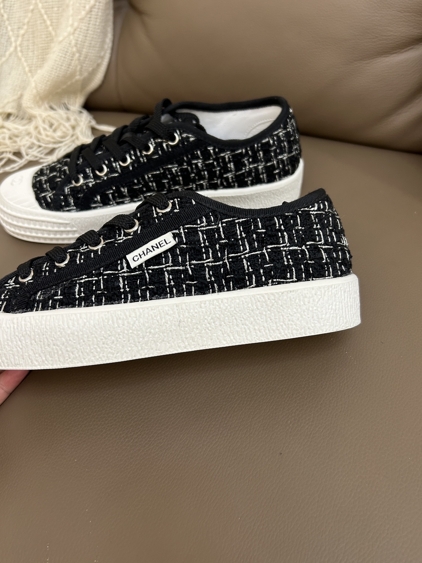 Pair of Chanel black and white tweed sneakers with white rubber toe caps featuring the CC logo on a brown leather surface - Hình 8