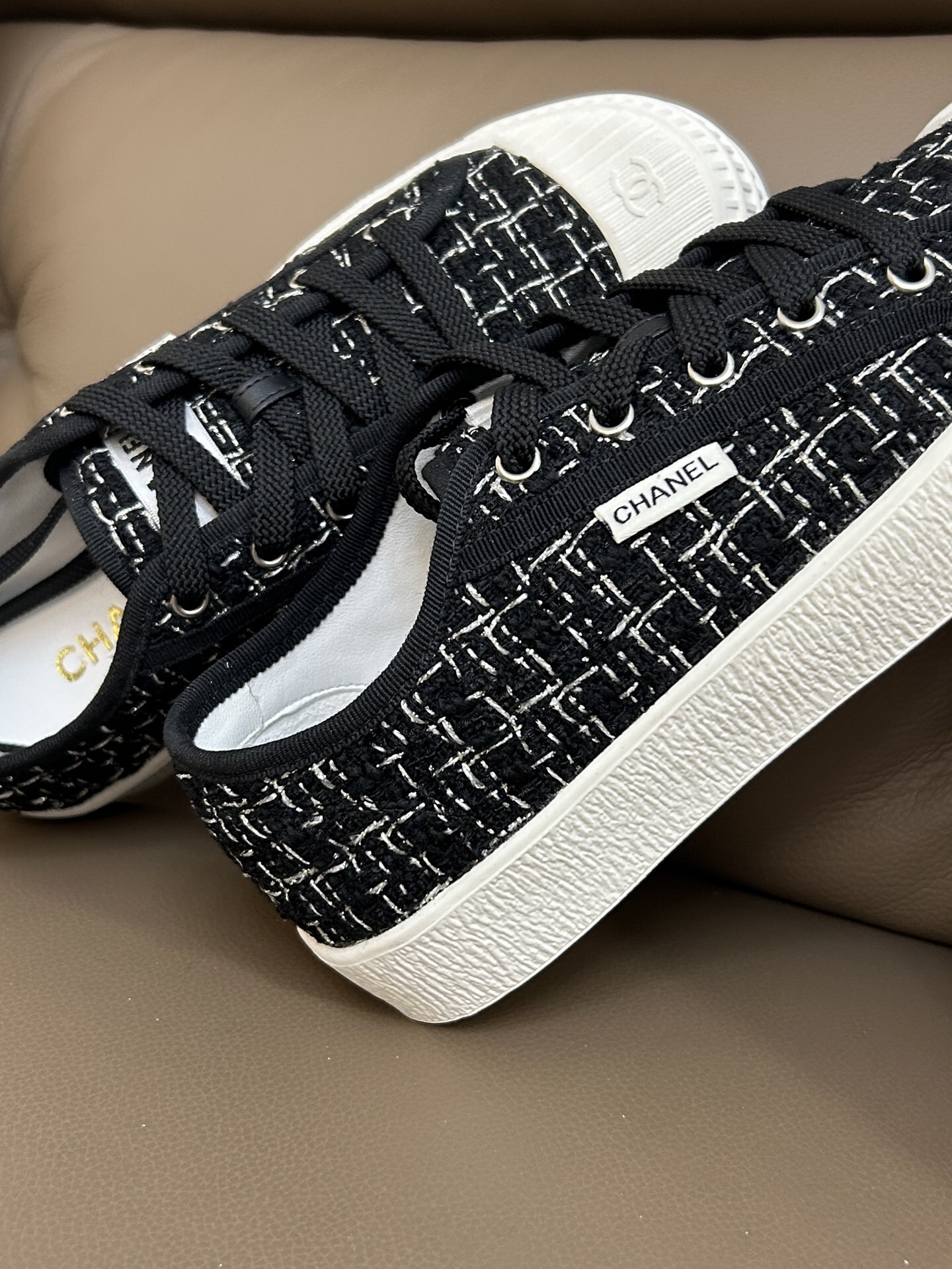 Pair of Chanel black and white tweed sneakers with white rubber toe caps featuring the CC logo on a brown leather surface - Hình 6