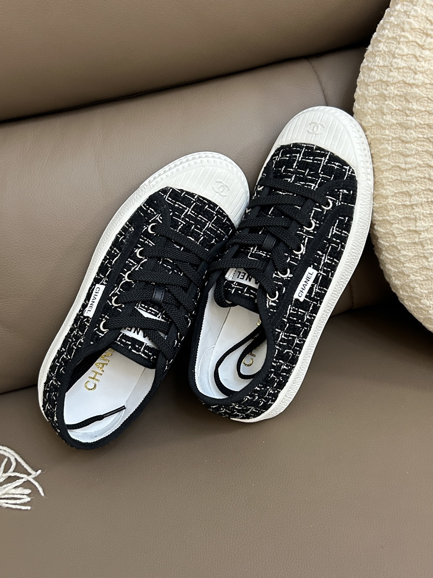 Pair of Chanel black and white tweed sneakers with white rubber toe caps featuring the CC logo on a brown leather surface - Hình 3