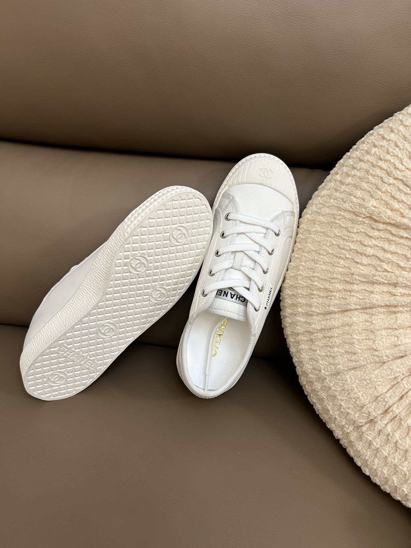 A pair of luxury Chanel white low-top sneakers with interlocking CC logo on the toe cap sitting on a brown leather surface. - Hình 8