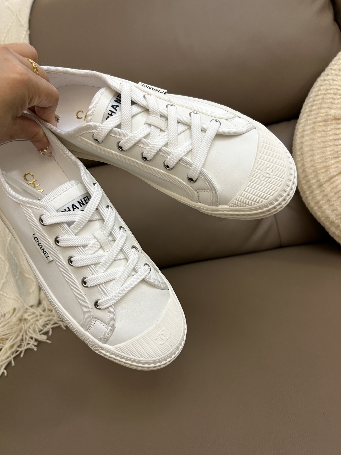 A pair of luxury Chanel white low-top sneakers with interlocking CC logo on the toe cap sitting on a brown leather surface. - Hình 6