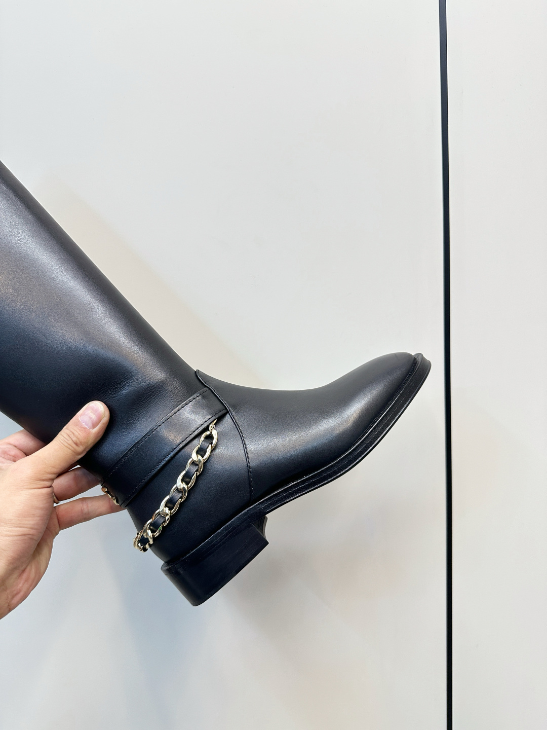 Pair of Chanel black leather knee-high boots with gold chain ankle detail on a wooden surface next to a Chanel shoe box. - Hình 7