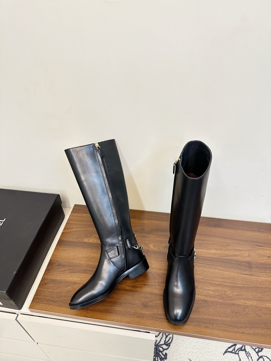 Pair of Chanel black leather knee-high boots with gold chain ankle detail on a wooden surface next to a Chanel shoe box. - Hình 5