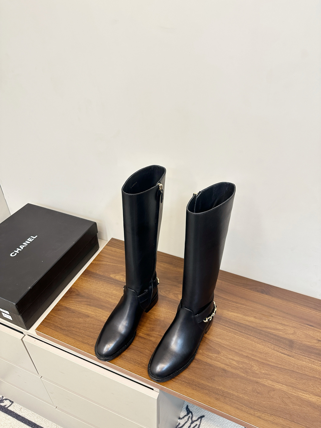 Pair of Chanel black leather knee-high boots with gold chain ankle detail on a wooden surface next to a Chanel shoe box. - Hình 3