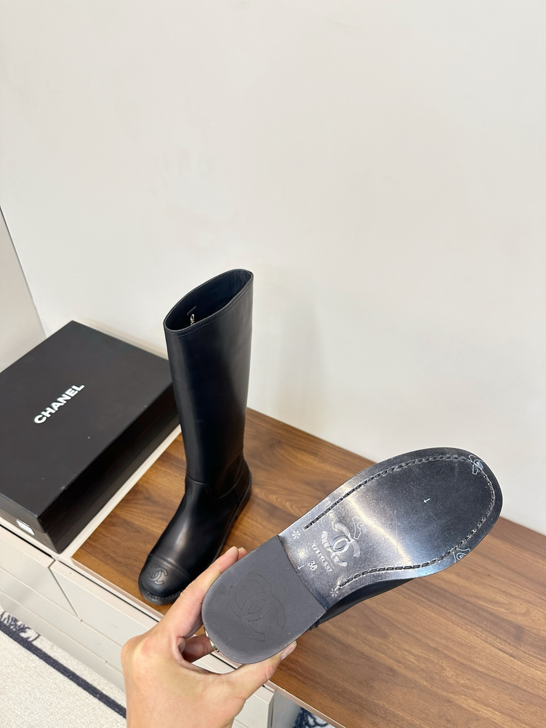 A pair of black Chanel knee-high leather riding boots featuring the embossed CC logo on the toe cap, displayed on a wooden surface next to a Chanel shoebox. - Hình 9