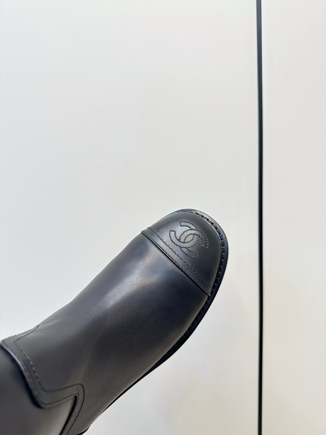 A pair of black Chanel knee-high leather riding boots featuring the embossed CC logo on the toe cap, displayed on a wooden surface next to a Chanel shoebox. - Hình 7