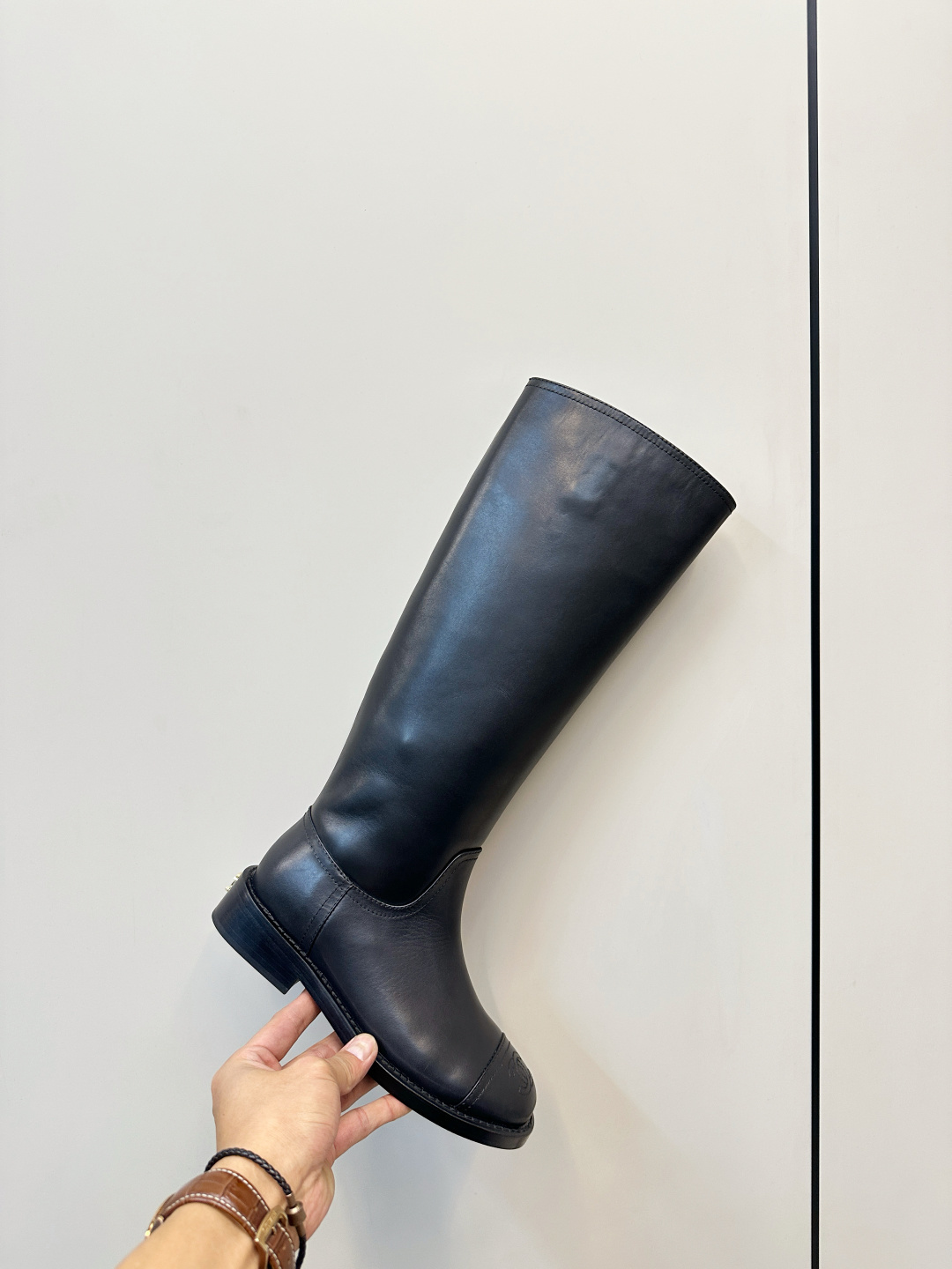 A pair of black Chanel knee-high leather riding boots featuring the embossed CC logo on the toe cap, displayed on a wooden surface next to a Chanel shoebox. - Hình 6