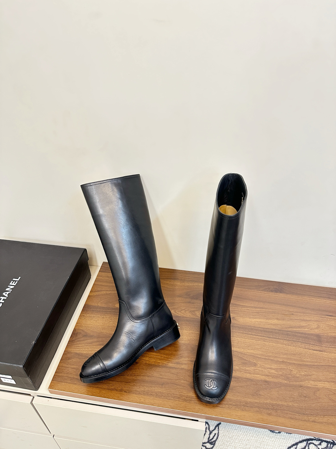 A pair of black Chanel knee-high leather riding boots featuring the embossed CC logo on the toe cap, displayed on a wooden surface next to a Chanel shoebox. - Hình 5