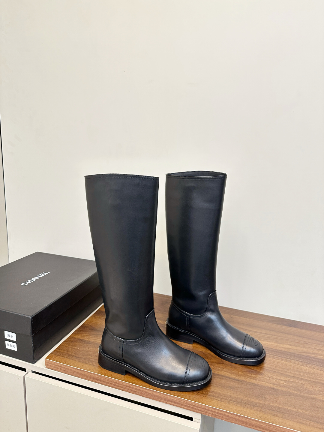 A pair of black Chanel knee-high leather riding boots featuring the embossed CC logo on the toe cap, displayed on a wooden surface next to a Chanel shoebox. - Hình 2