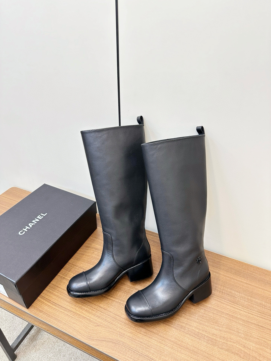 Pair of Chanel black leather knee-high riding boots with block heels and cap toes sitting on a wooden bench next to a Chanel shoe box. - Hình 9