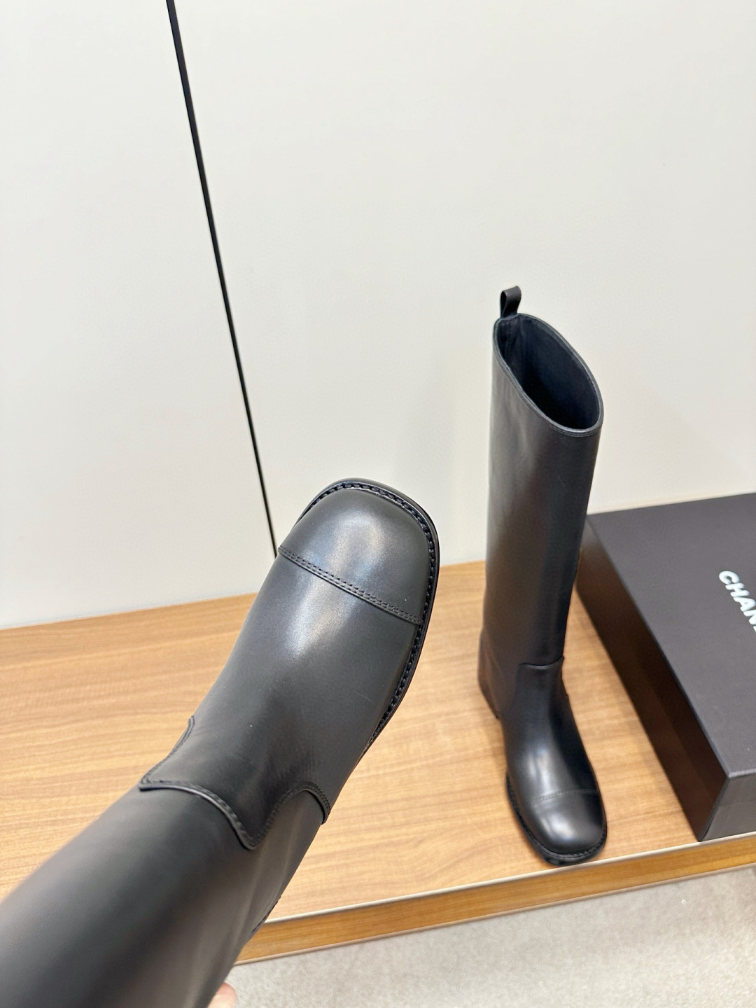 Pair of Chanel black leather knee-high riding boots with block heels and cap toes sitting on a wooden bench next to a Chanel shoe box. - Hình 8