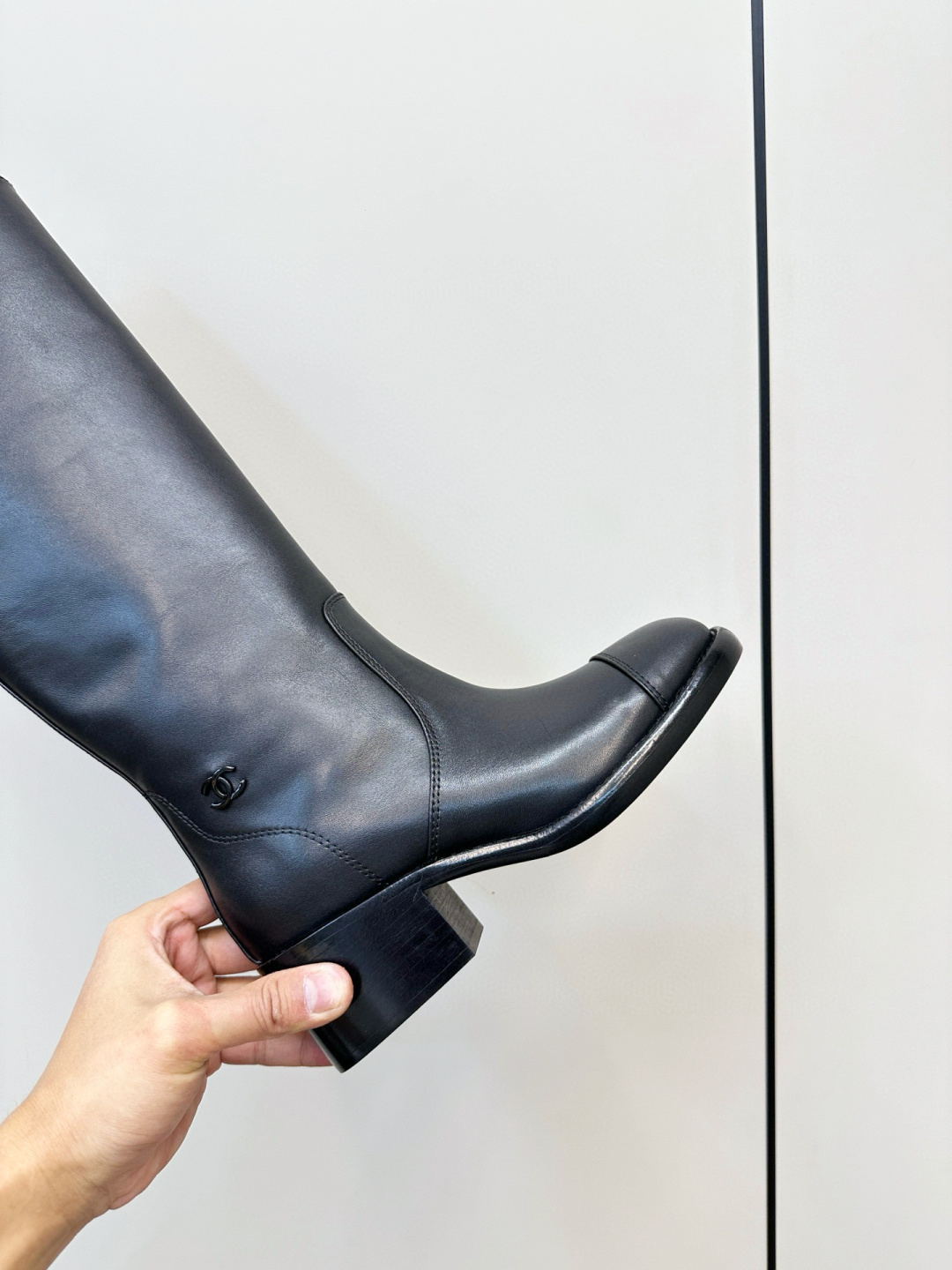 Pair of Chanel black leather knee-high riding boots with block heels and cap toes sitting on a wooden bench next to a Chanel shoe box. - Hình 7