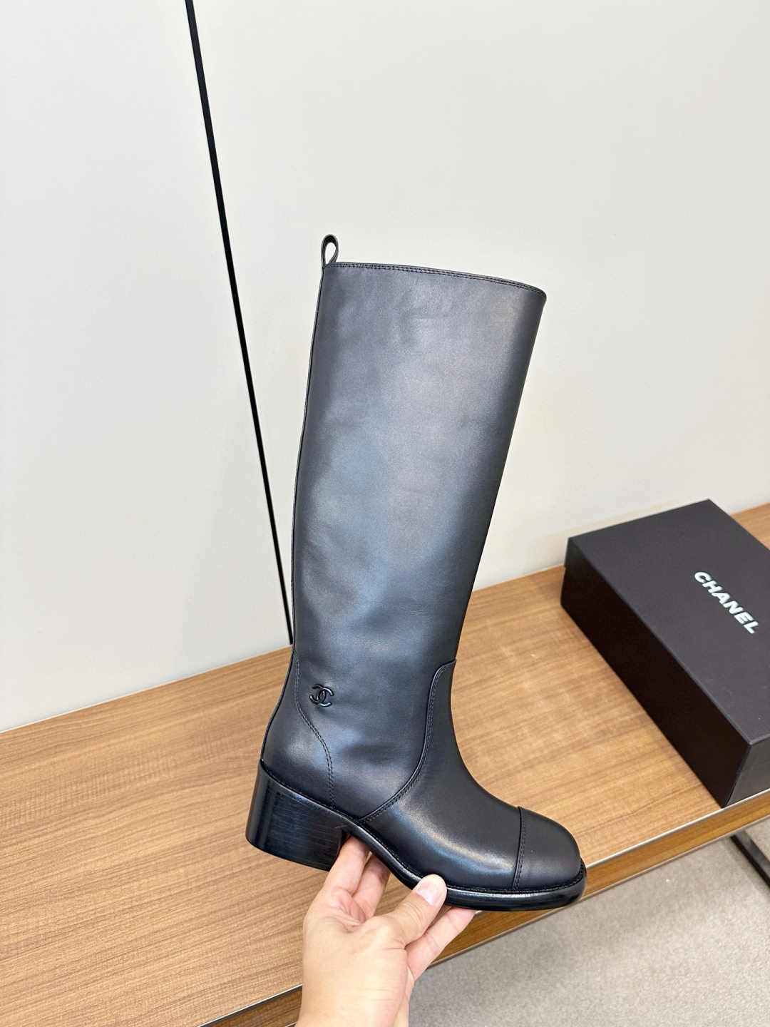 Pair of Chanel black leather knee-high riding boots with block heels and cap toes sitting on a wooden bench next to a Chanel shoe box. - Hình 6