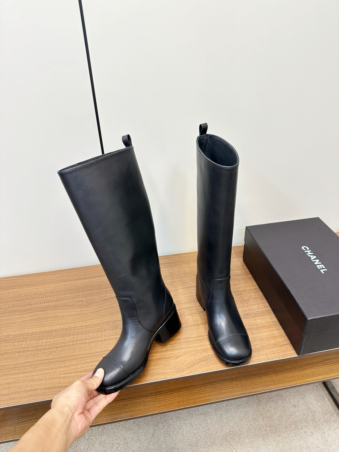 Pair of Chanel black leather knee-high riding boots with block heels and cap toes sitting on a wooden bench next to a Chanel shoe box. - Hình 5