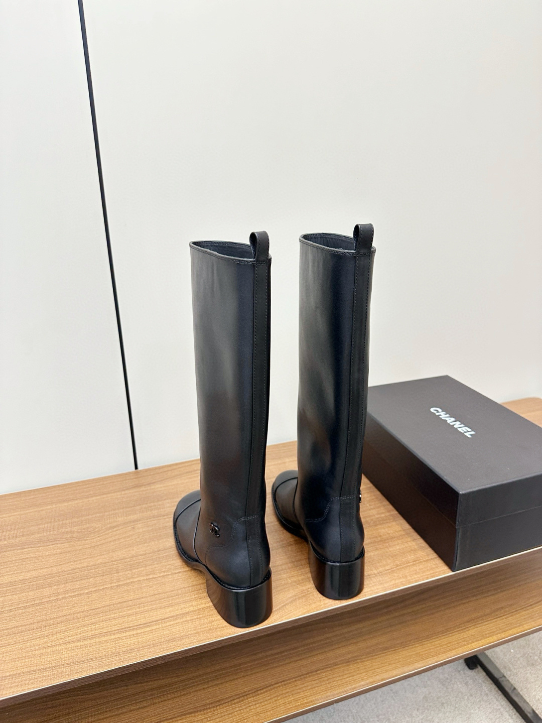 Pair of Chanel black leather knee-high riding boots with block heels and cap toes sitting on a wooden bench next to a Chanel shoe box. - Hình 4
