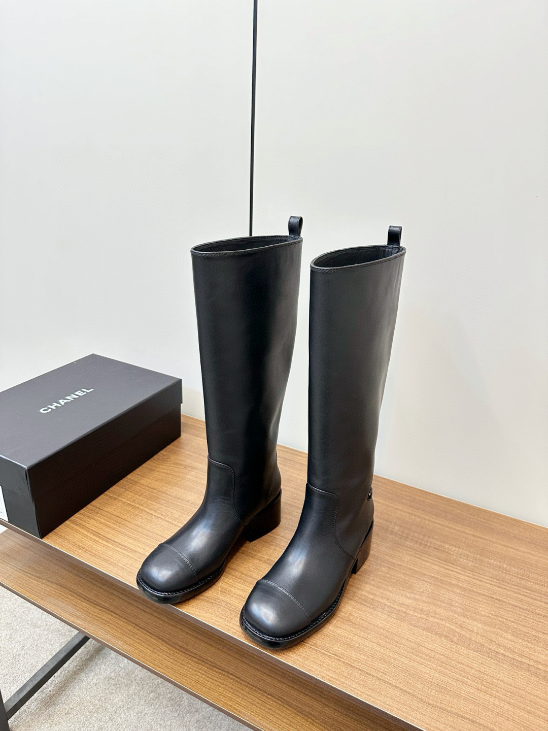 Pair of Chanel black leather knee-high riding boots with block heels and cap toes sitting on a wooden bench next to a Chanel shoe box. - Hình 3