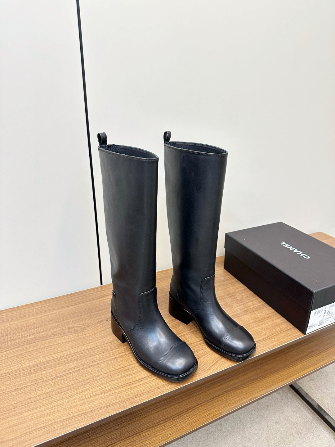Pair of Chanel black leather knee-high riding boots with block heels and cap toes sitting on a wooden bench next to a Chanel shoe box. - Hình 2