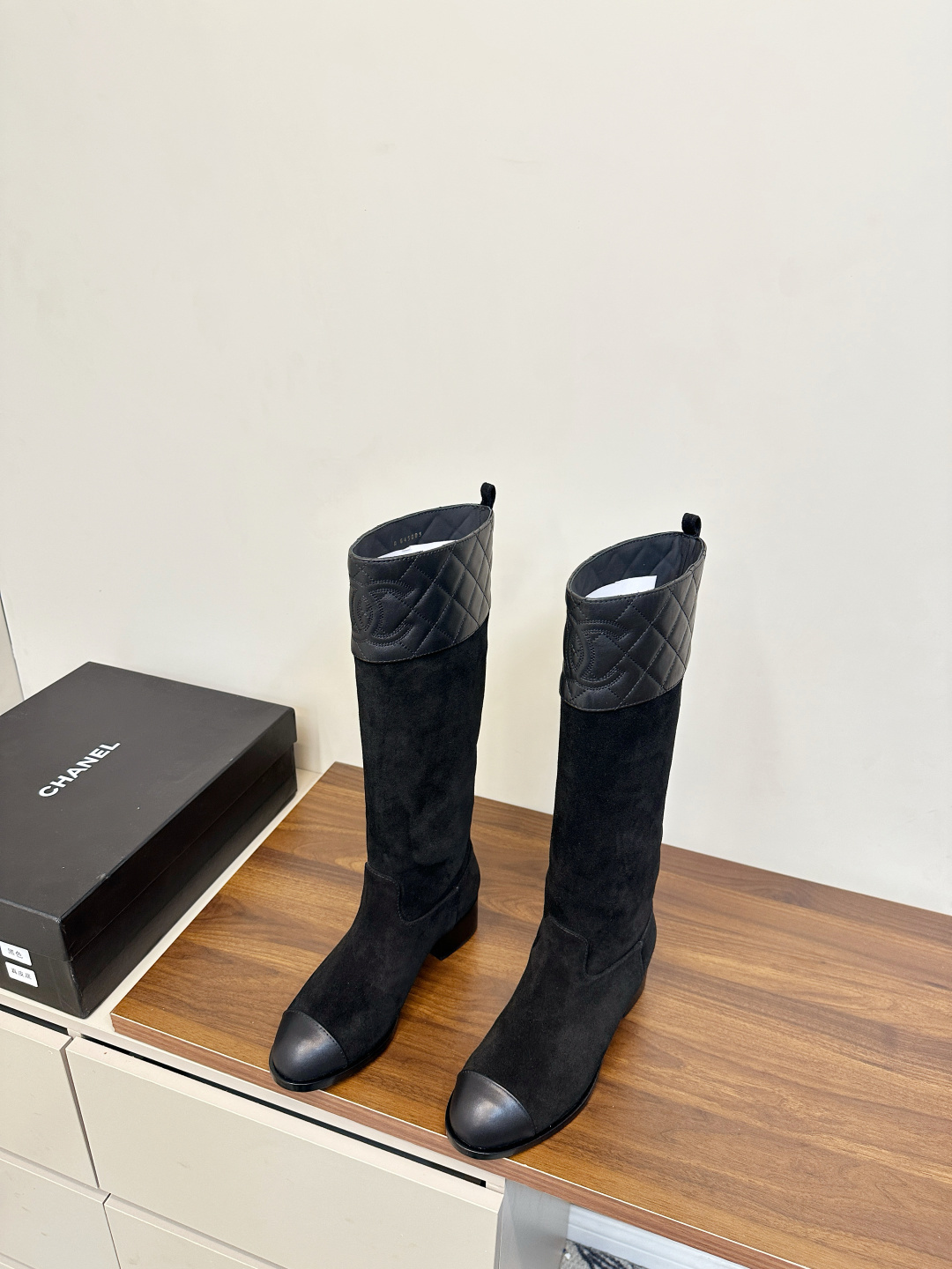 A pair of Chanel black suede knee-high boots with quilted leather tops and leather toe caps on a wooden surface. - Hình 3