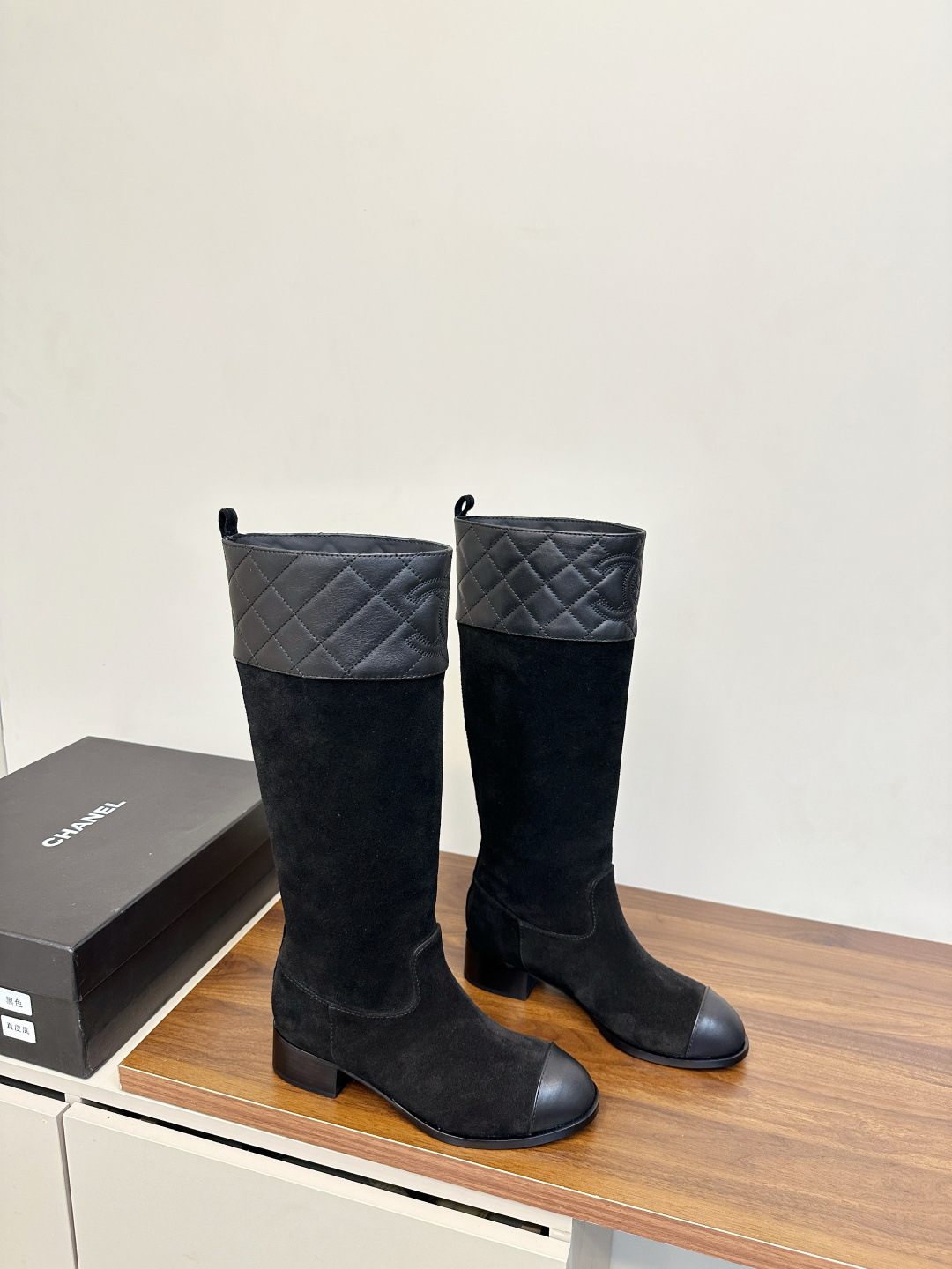 A pair of Chanel black suede knee-high boots with quilted leather tops and leather toe caps on a wooden surface. - Hình 2
