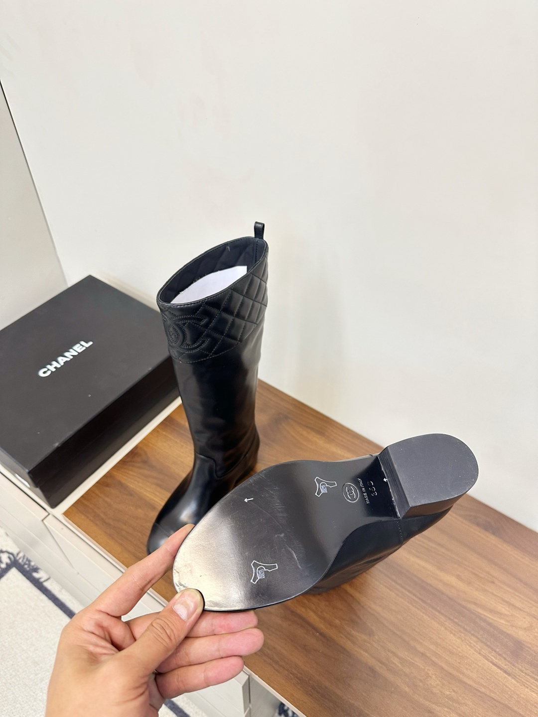 A pair of black Chanel knee-high leather boots with quilted tops and stitched CC logo displayed on a wooden surface. - Hình 9