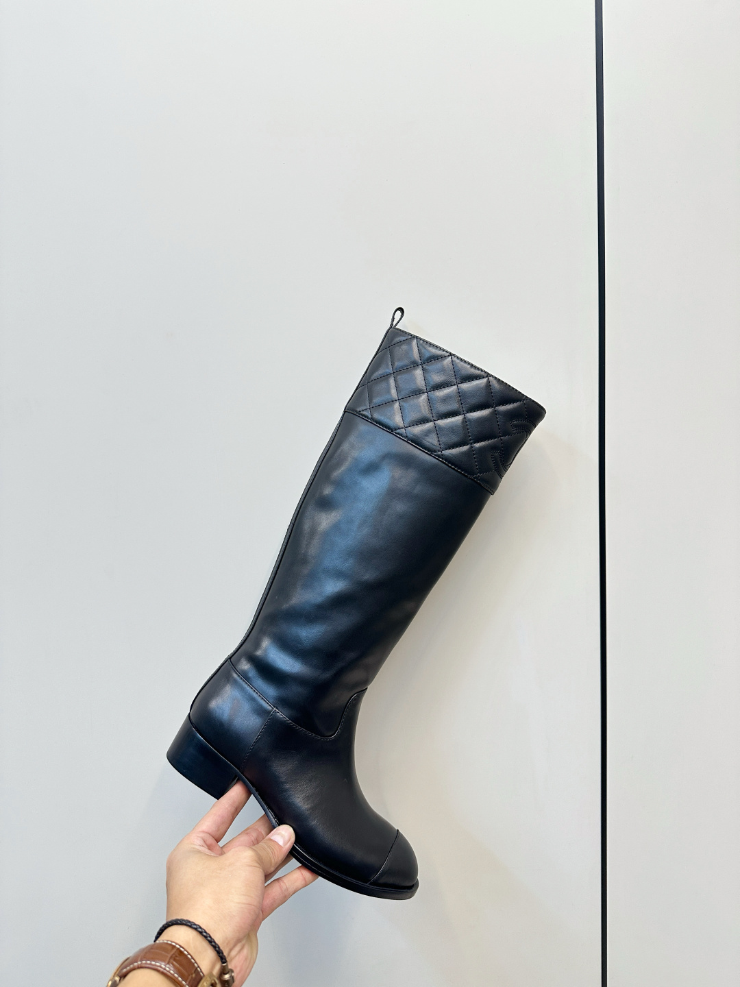 A pair of black Chanel knee-high leather boots with quilted tops and stitched CC logo displayed on a wooden surface. - Hình 6