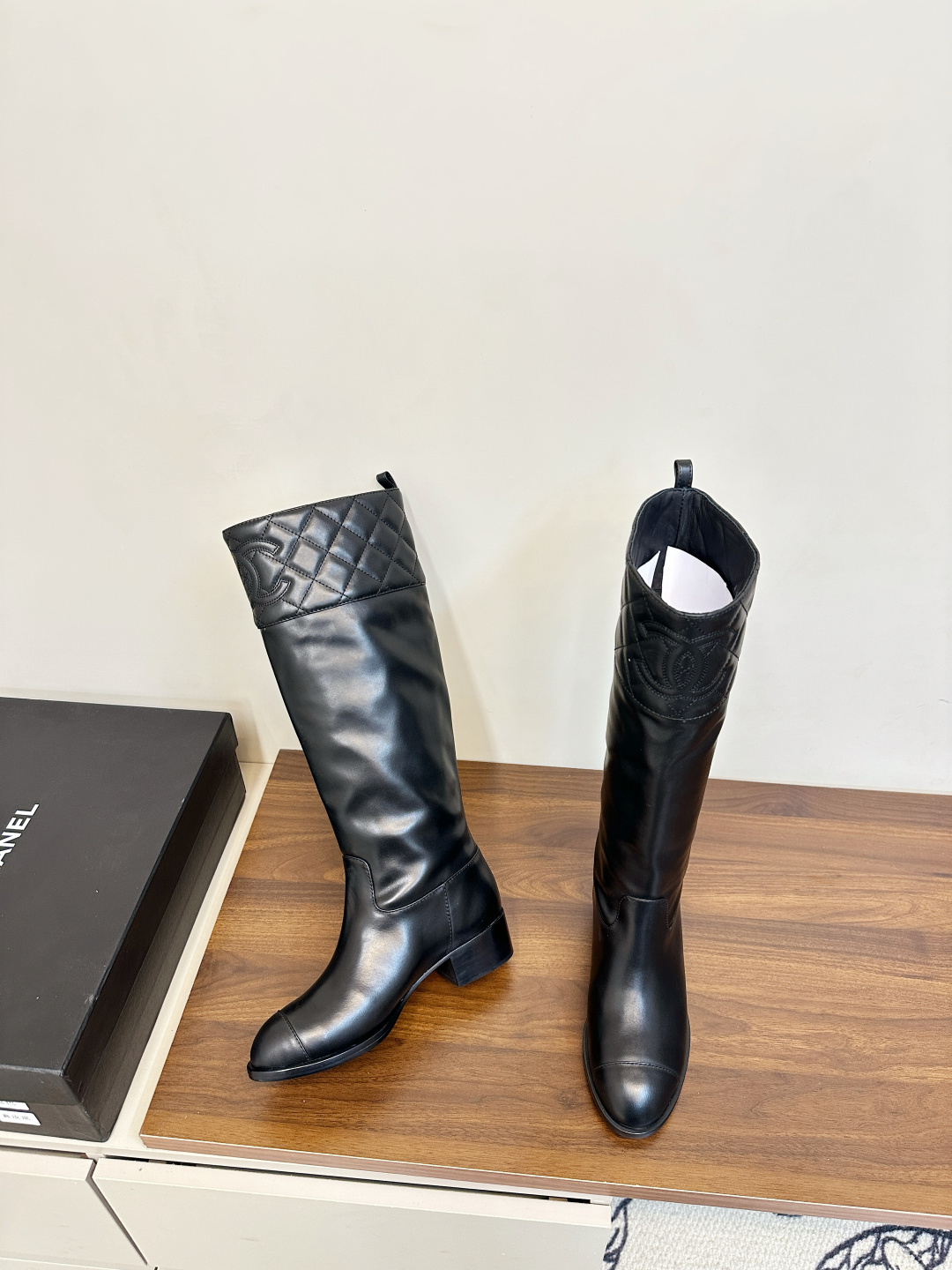 A pair of black Chanel knee-high leather boots with quilted tops and stitched CC logo displayed on a wooden surface. - Hình 5