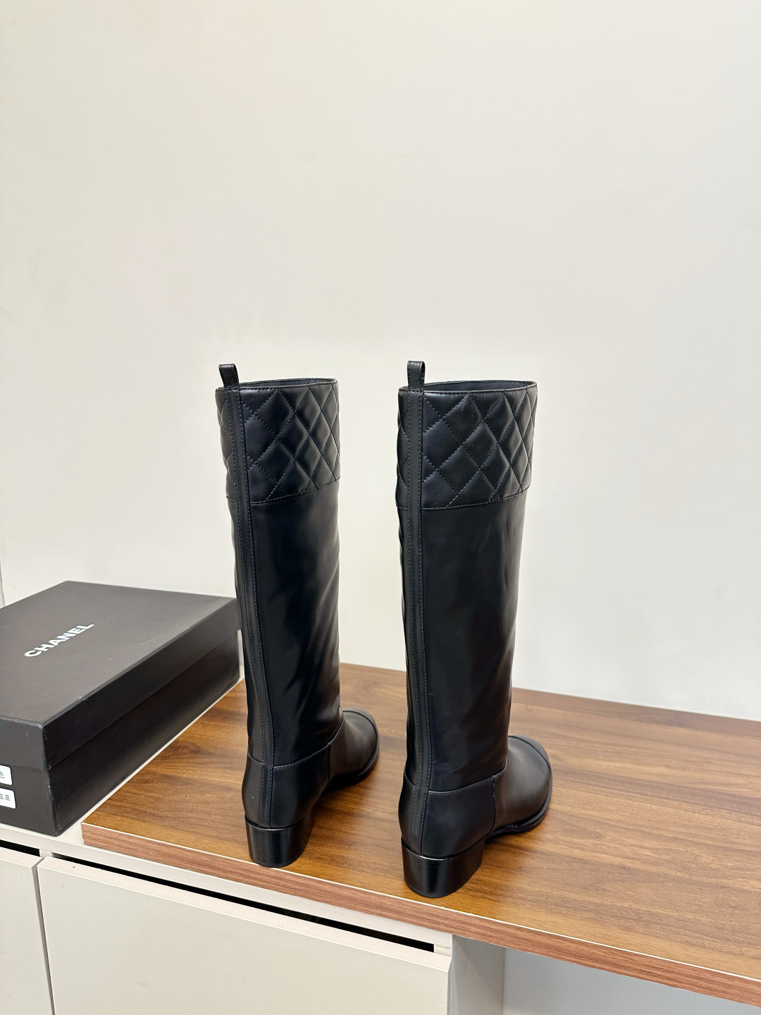 A pair of black Chanel knee-high leather boots with quilted tops and stitched CC logo displayed on a wooden surface. - Hình 4