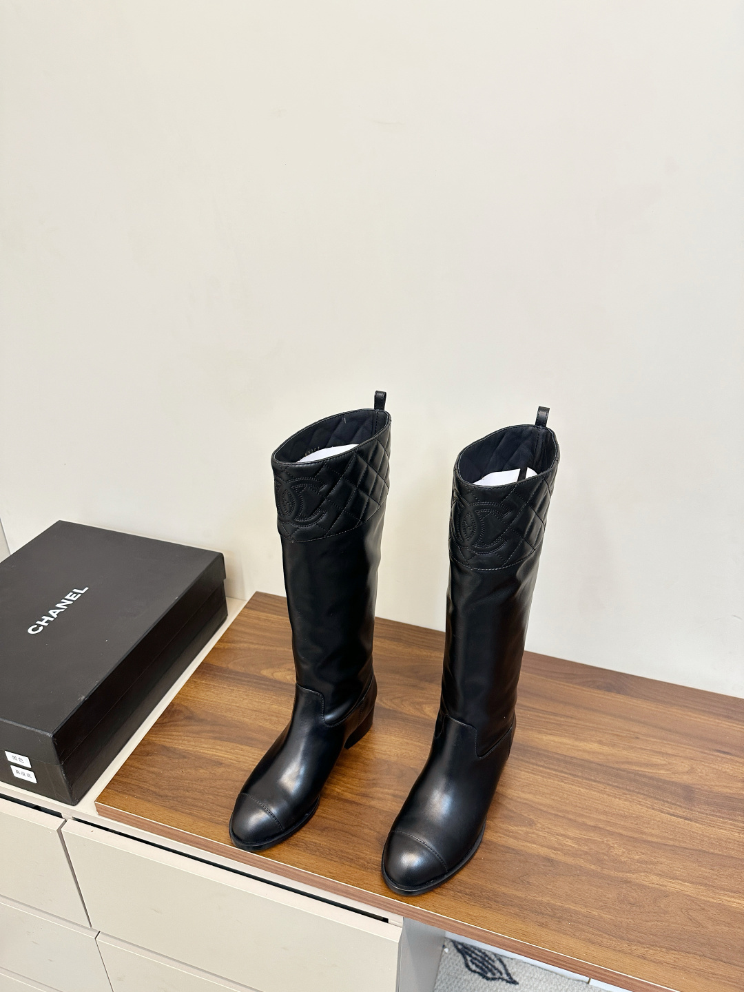 A pair of black Chanel knee-high leather boots with quilted tops and stitched CC logo displayed on a wooden surface. - Hình 3
