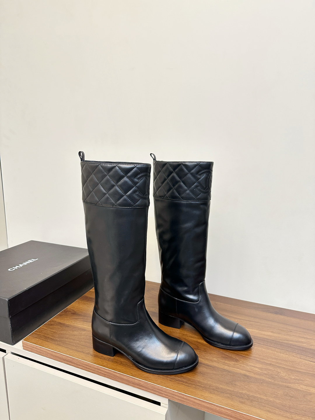 A pair of black Chanel knee-high leather boots with quilted tops and stitched CC logo displayed on a wooden surface. - Hình 2