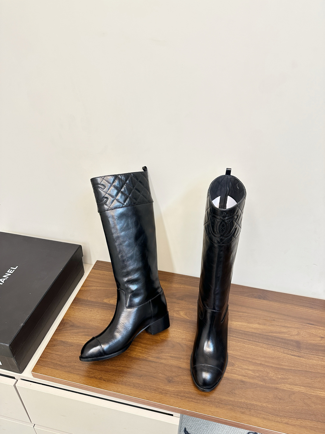 Pair of Chanel black quilted leather knee-high boots with embossed CC logo on a wooden surface. - Hình 5