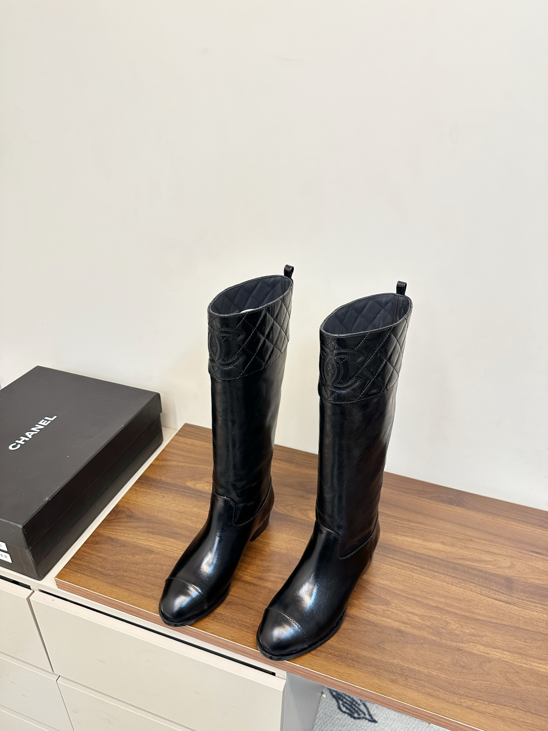 Pair of Chanel black quilted leather knee-high boots with embossed CC logo on a wooden surface. - Hình 3