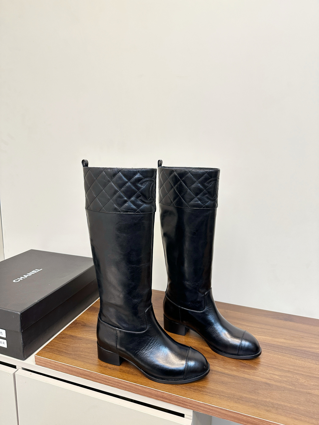 Pair of Chanel black quilted leather knee-high boots with embossed CC logo on a wooden surface. - Hình 2