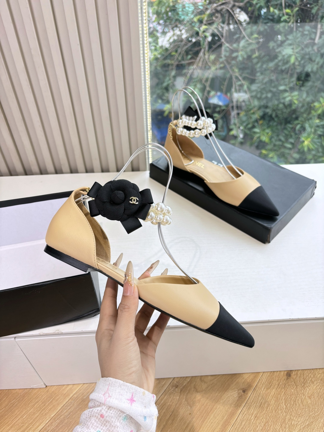 Elegant beige pointed-toe flats with black tips, featuring double-layered pearl ankle straps and a black camellia flower accent. - Hình 3