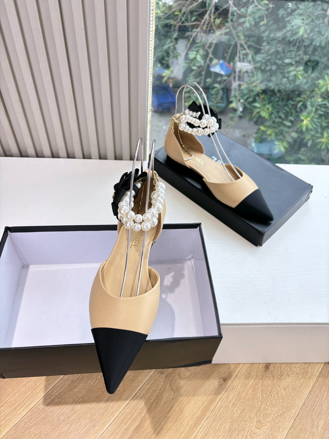 Elegant beige pointed-toe flats with black tips, featuring double-layered pearl ankle straps and a black camellia flower accent. - Hình 2