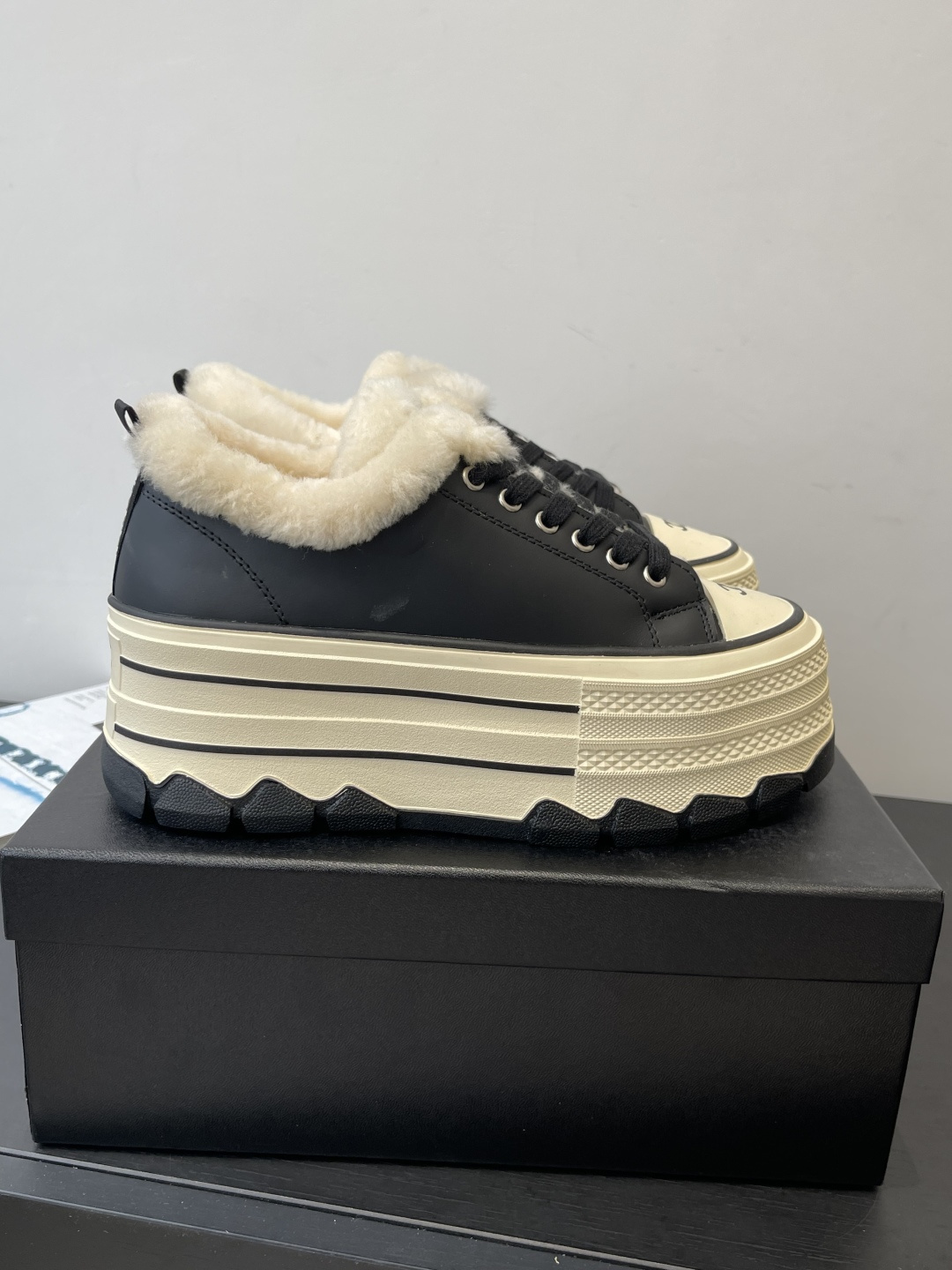 A pair of Chanel black leather platform sneakers with cream shearling lining and iconic CC logo on the toe cap, sitting on a black Chanel shoe box. - Hình 2