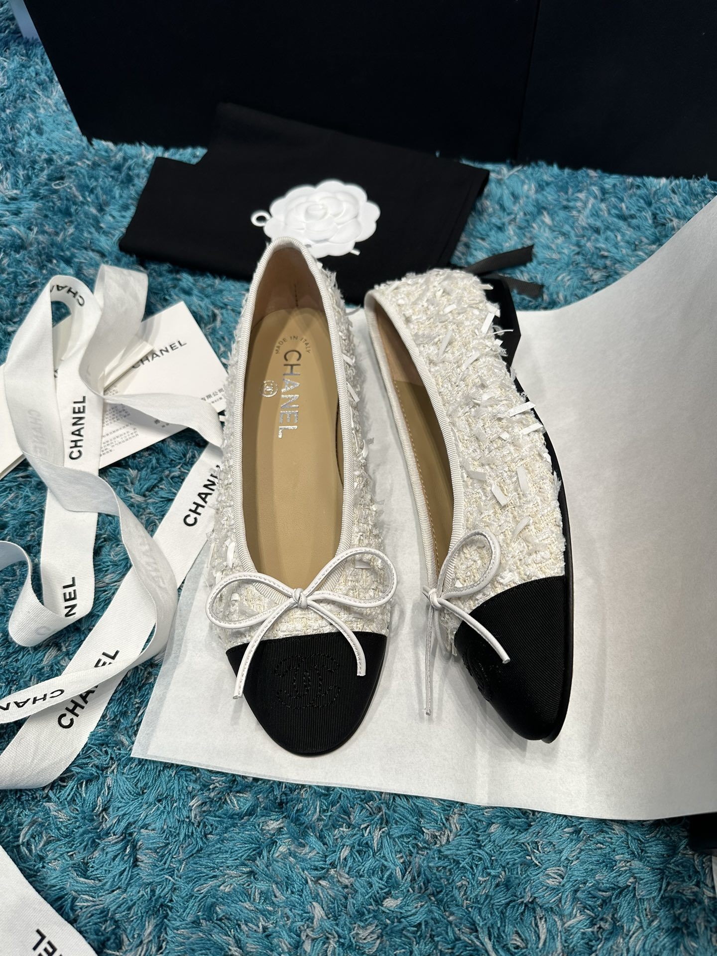A pair of Chanel white tweed ballet flats with black toe caps and white bows, presented in a Chanel box with branded ribbons. - Hình 2