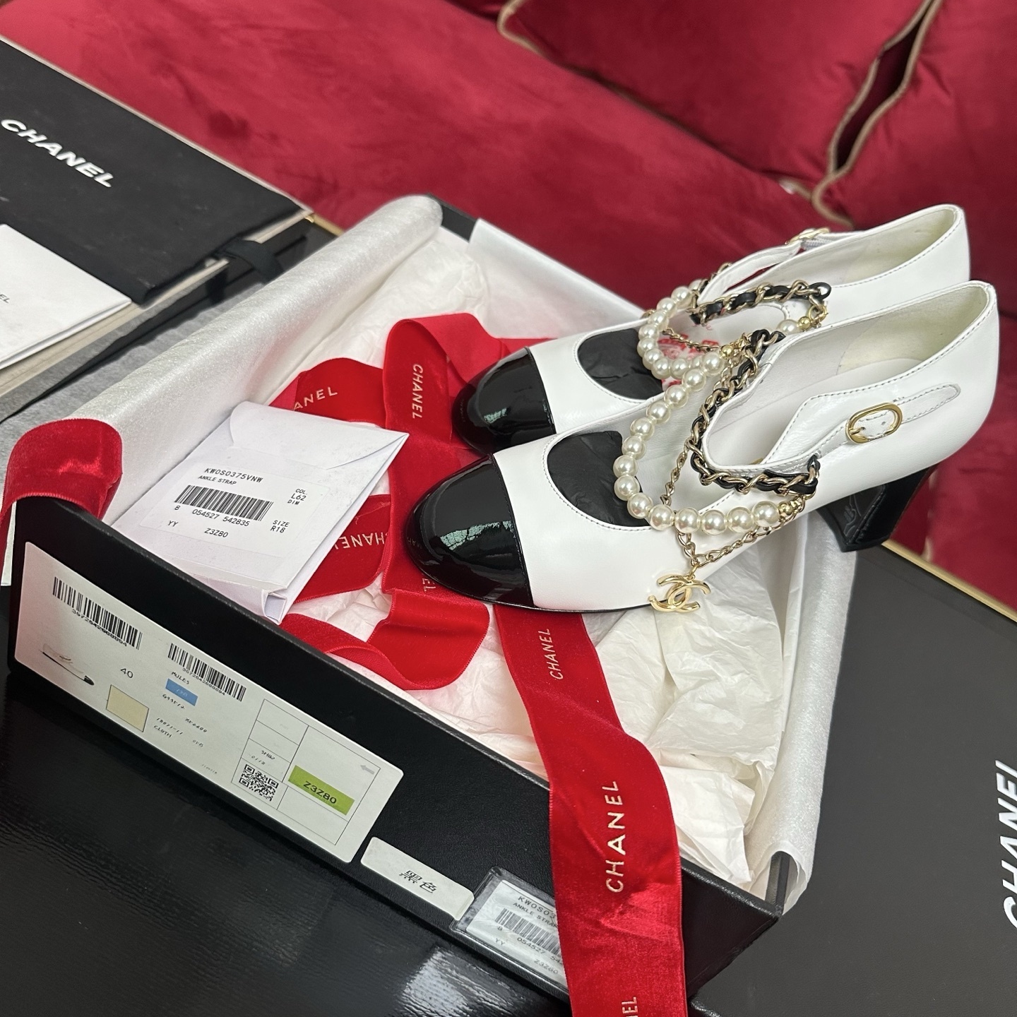Pair of white Chanel Mary Jane pumps with black patent leather cap toes and decorative pearl and gold chain ankle straps in a luxury box. - Hình 2