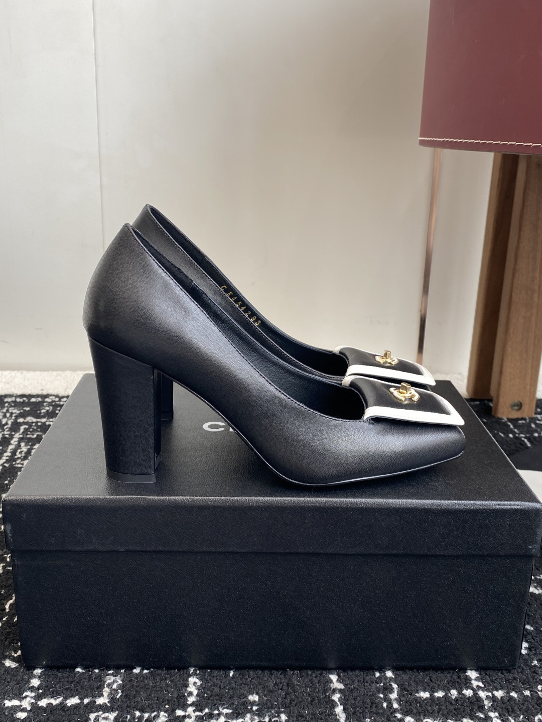 Chanel black leather square toe pumps with gold CC logo and white trim detail on a luxury shoe box - Hình 2