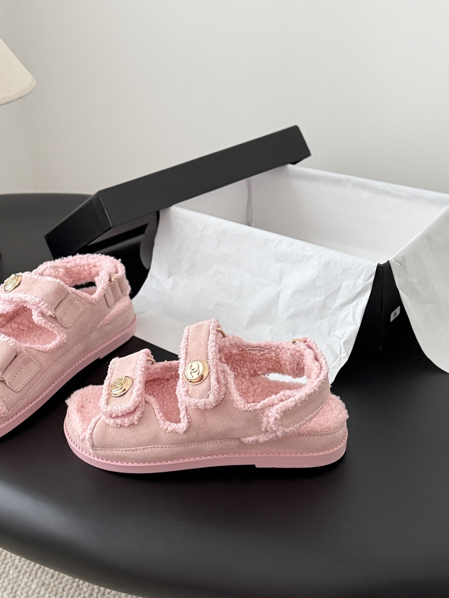 Pair of pink Chanel shearling sandals with gold CC logo buttons sitting next to a black Chanel box. - Hình 2