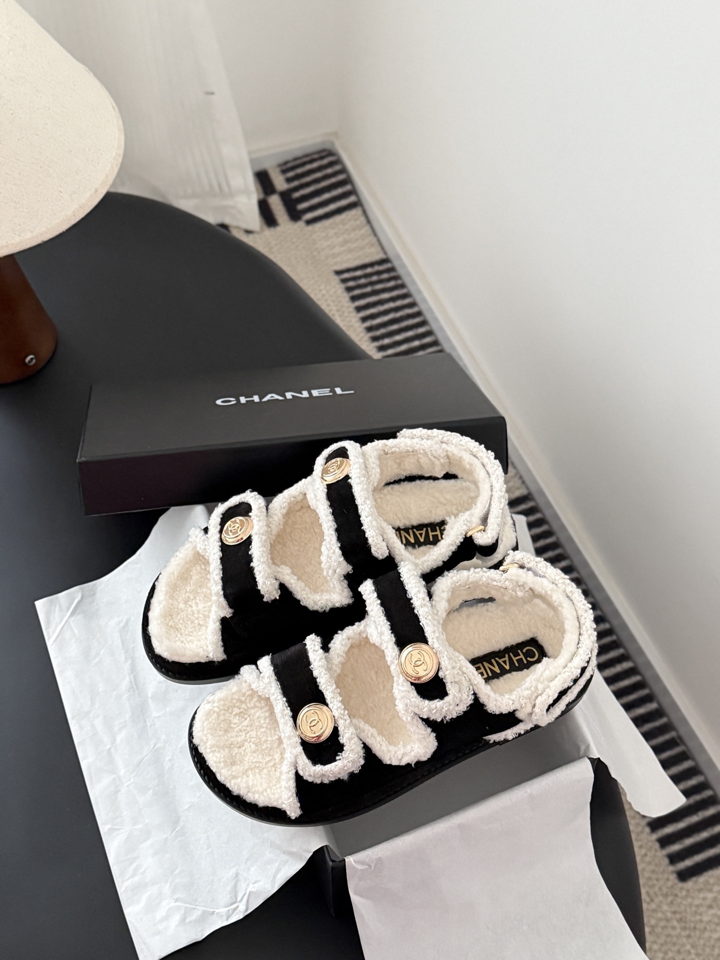 Chanel black and white shearling dad sandals with gold CC logo buttons on a black table next to a Chanel box - Hình 9